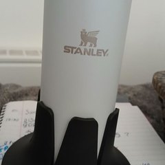 A white Stanley insulated tumbler seated in a black drink base standing on an open spiral planner covered in handwritten to-do notes, with a basket of pens alongside