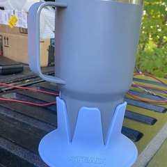 A grey insulated tumbler with a handle in a periwinkle STEADI base standing on the black plastic bed rail of a pickup truck, cargo and wiring in the bed behind, green trees beyond